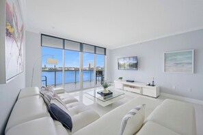 Un salon moderne doté d’une grande fenêtre, d’un ensemble de canapés blancs, d’une table basse en verre et d’une console blanche sur laquelle est posée une télévision à écran plat.