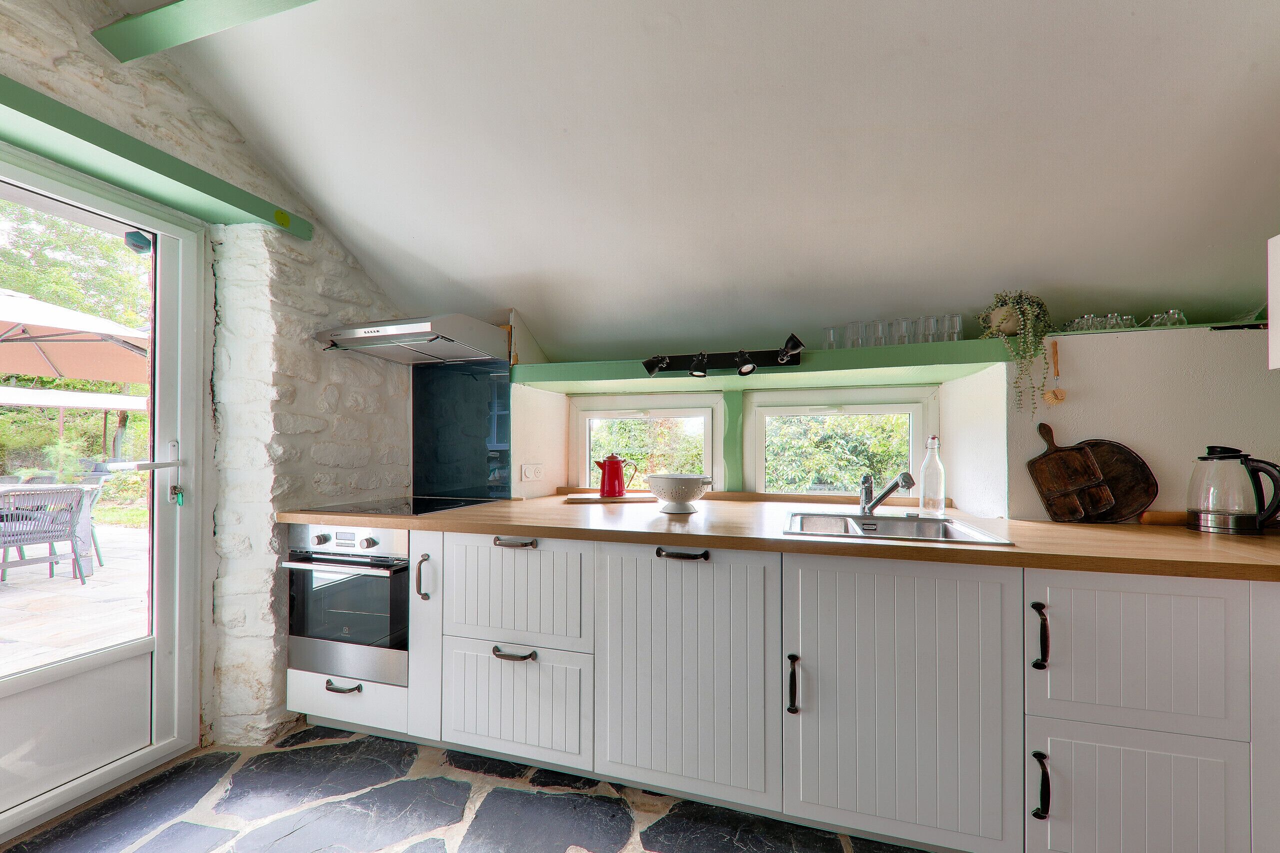 Una cucina con mobili bianchi, pavimento in pietra e finestra con vista su un giardino.