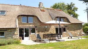 Une maison en pierre avec un toit de chaume, une terrasse avec des meubles d’extérieur et un grand parasol.
