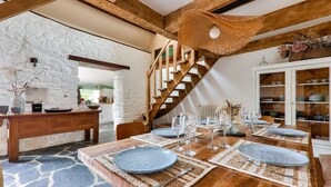 Un coin repas avec une table en bois dressée pour six personnes, un mur en pierre et un escalier avec des poutres en bois.