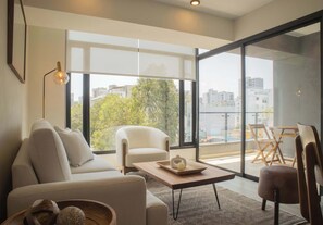 Una sala de estar moderna con sofá, sillón y mesa de centro, ventanas amplias con vista a la ciudad y un balcón con mobiliario exterior.