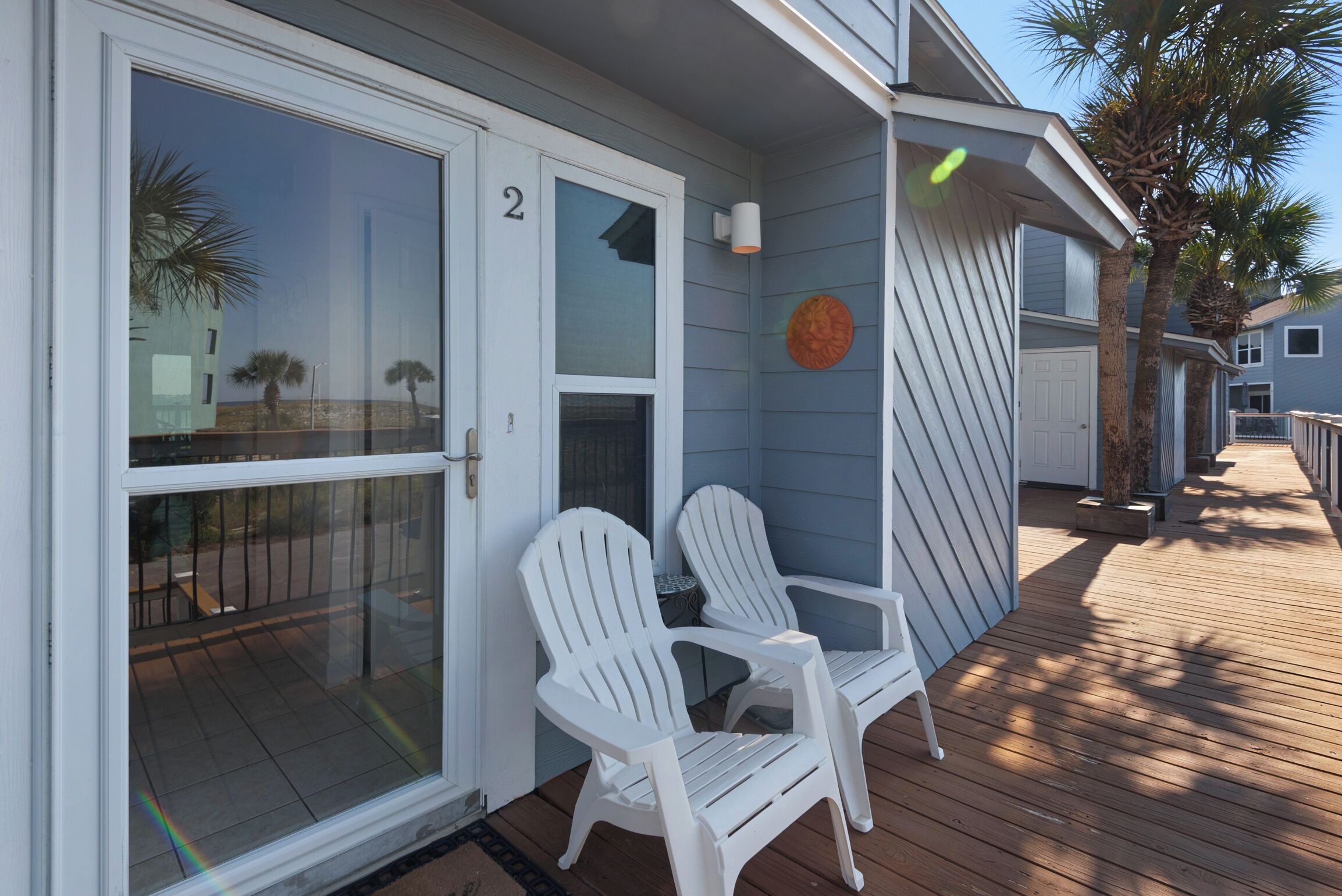 A wooden deck with two white Adirondack chairs, a glass door with a number '2', and a palm tree.