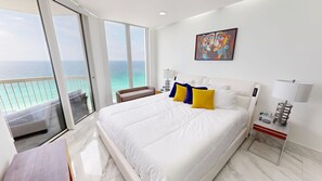 Habitación de hotel con una cama grande, un balcón con vista al océano, un cuadro en la pared y una mesita de noche con una lámpara.