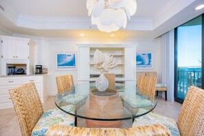 Un comedor con una mesa de vidrio rodeada de sillas de mimbre, muebles de cocina blancos y obras de arte en las paredes.