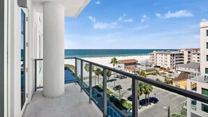 Un balcon avec vue sur la plage et l’océan, un parking et un immeuble à plusieurs étages.
