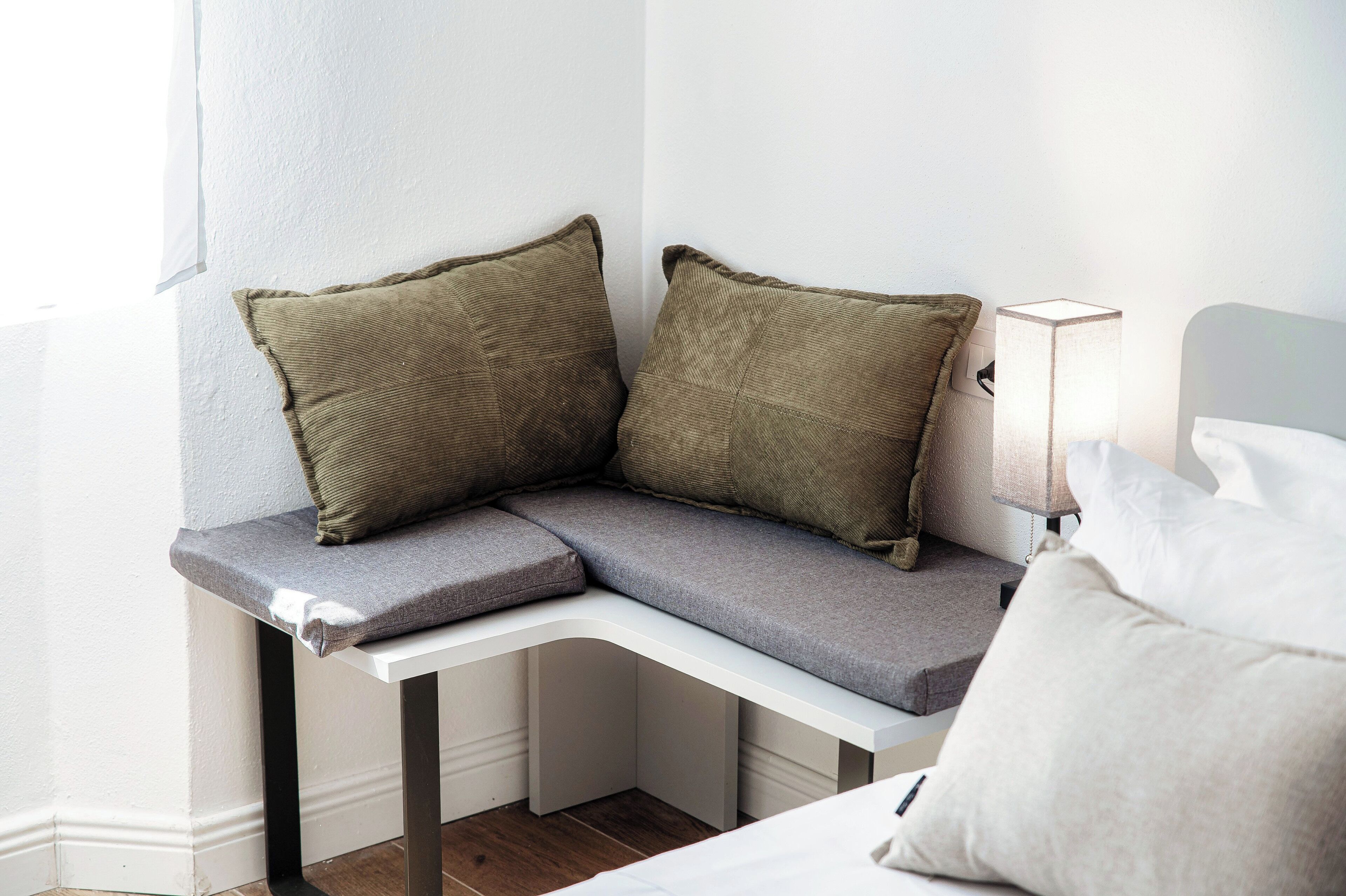 Een zithoek met een bank en kussens, een nachtkastje met een lamp, en een bed met kussens.