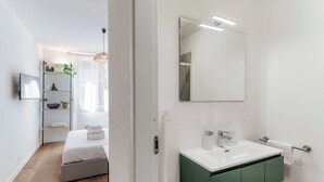 A modern bedroom with a bed, a bedside table, a mirror, and a bathroom with a sink and green cabinets. - Batèl House 1 - By Breva Collections (Como)