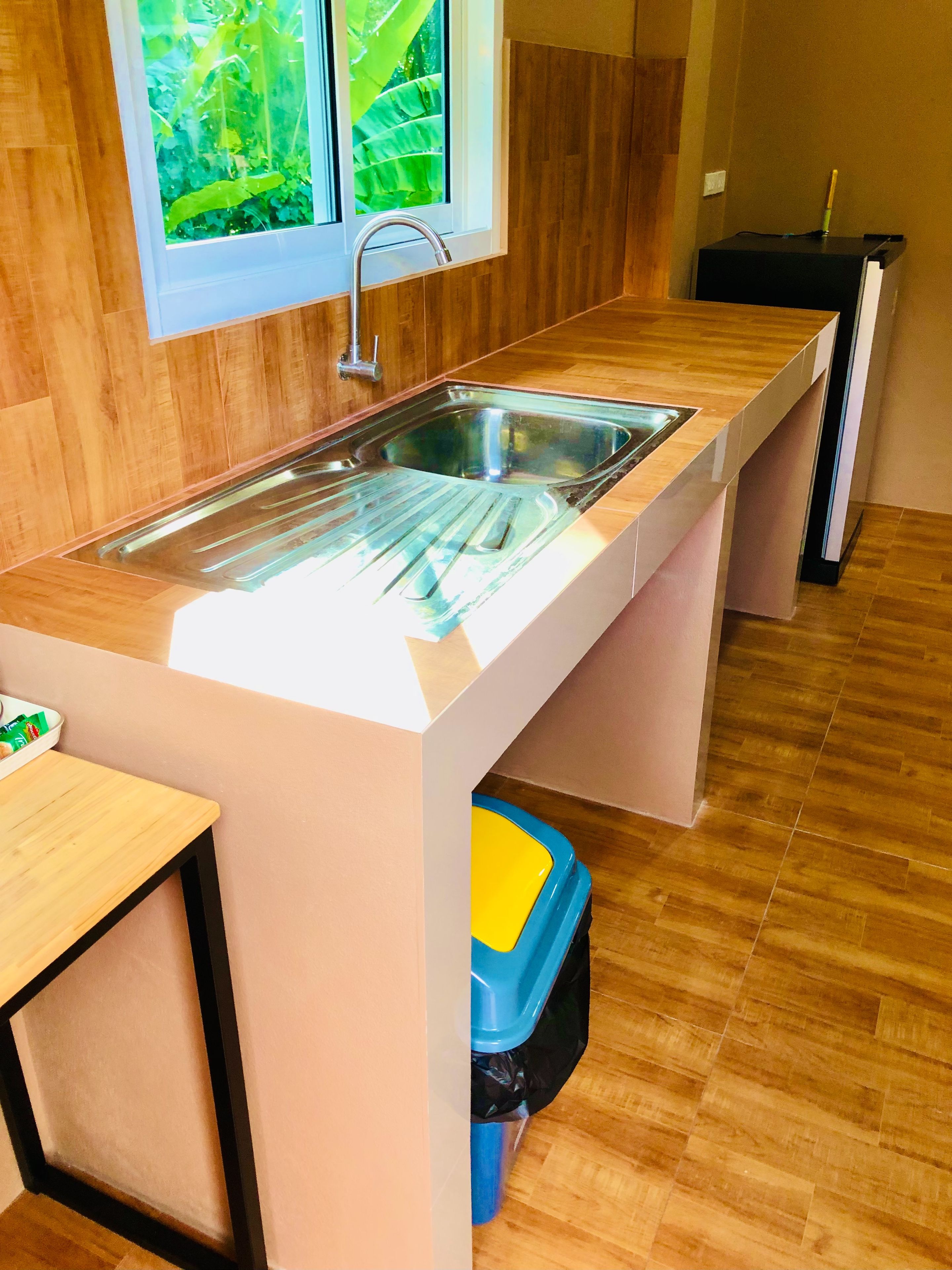 A modern kitchen with a stainless steel sink, wooden flooring, and a window with a view of greenery.