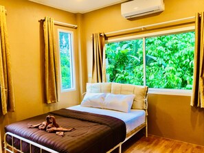 A bedroom with a bed, a window with curtains, an air conditioner, and a view of greenery outside. - Lucky Sky Homestay (Ko Pha-ngan)