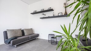 A modern living room with a grey sofa, two black shelves with framed pictures, a vase with dried plants, and a tall green plant.