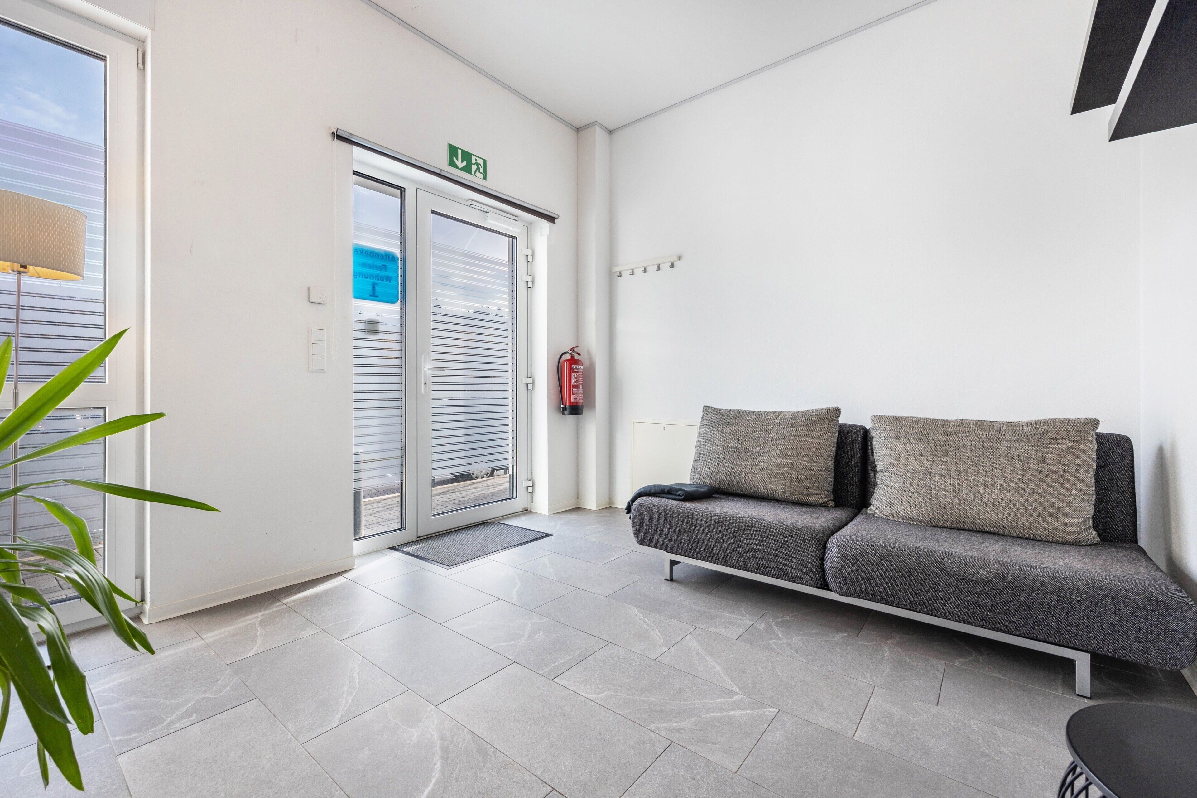 A reception area with a sofa, a fire extinguisher, a door with an emergency exit sign, and a plant.