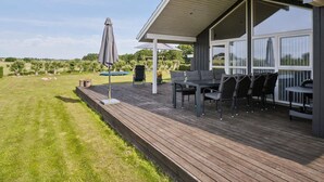 A covered outdoor patio with a table and chairs, a swing set, and a view of a grassy field and trees.