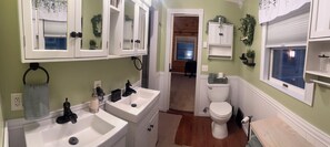 Une salle de bain avec deux lavabos, des toilettes et une fenêtre. Il y a des armoires au-dessus des lavabos et une petite étagère au-dessus des toilettes.