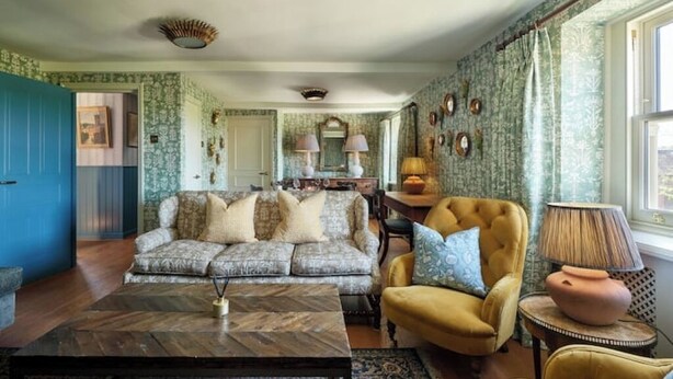 A living room with a sofa, armchair, and coffee table. There is a door on the left, a window on the right, and a lamp on a side table.