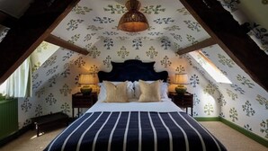 A bedroom with a large bed, two bedside tables with lamps, a hanging light fixture, and a sloped ceiling with floral wallpaper.