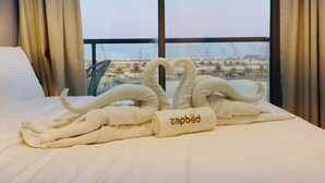 A neatly made bed with a towel shaped like a swan and a towel with "zapbed" embroidered on it, set against a backdrop of a large window with a view of a cityscape and construction cranes. - RAK F 211 (Ras Al Khaimah)