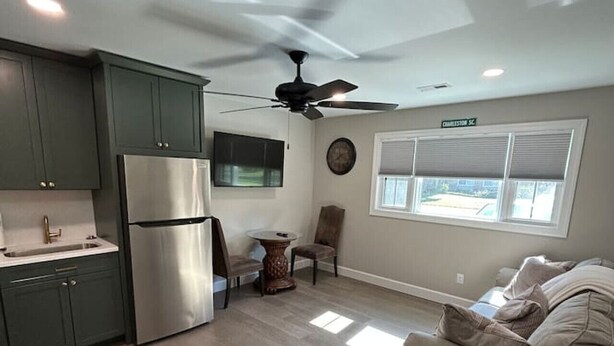 A modern kitchen with dark cabinets, a stainless steel refrigerator, and a ceiling fan. There is a dining area with a table and chairs, and a large window providing natural light.