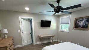 A bedroom with a bed, a gray armchair, a desk, a television, and a ceiling fan.