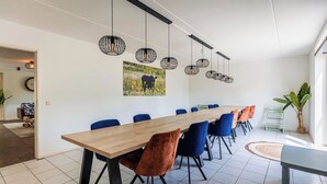 A modern dining area with a long wooden table, blue and orange chairs, hanging pendant lights, and a painting of a cow in a field. - Detached Bungalow by the Forest Edge (Ewijk)