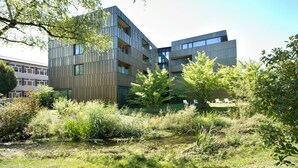 A modern building with multiple floors, surrounded by lush greenery and a small water feature. - Hotel am See (Hard)