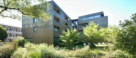 A modern building with multiple floors, surrounded by lush greenery and a small water feature.