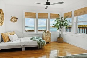 A room with a bed, a wooden floor, a ceiling fan, and large windows with a view of the sea.