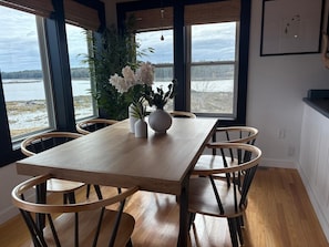 A dining area with a wooden table and chairs, a vase with flowers, and large windows with a view of a body of water.