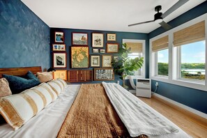 Ein Schlafzimmer mit einem Bett, einem großen Fenster, einem Deckenventilator und einer Wand mit gerahmten Bildern.
