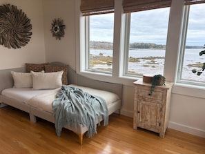A cozy room with a sofa, a small wooden cabinet, and a large window offering a view of a rocky shoreline.
