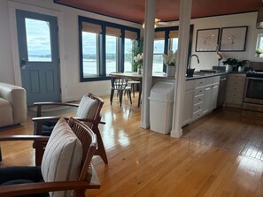 A spacious living area with a dining table, chairs, a kitchen with a stove, and large windows offering a view of the outdoors.