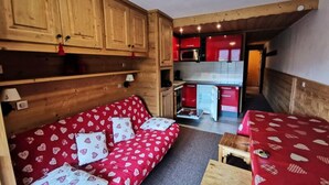 A compact living space with a wooden interior, a red sofa with heart patterns, and a kitchenette with red cabinets. - Apartment in Meribel With Ski Slope Views (Les Allues)