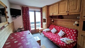 A room with a bed, a sofa, wooden cabinets, a television, and a window with curtains. - Apartment in Meribel With Ski Slope Views (Les Allues)