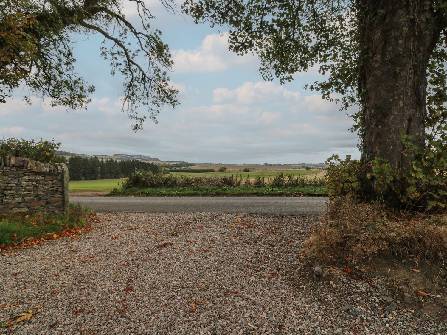 A gravel driveway leading to a stone wall with a rural landscape in the background.
