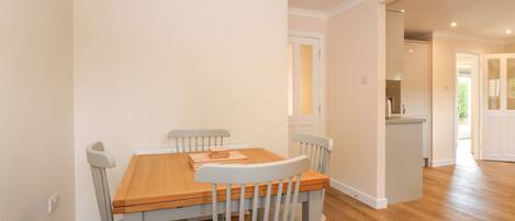 A dining area with a wooden table and four chairs, a kitchen in the background, and a hallway leading to another room.