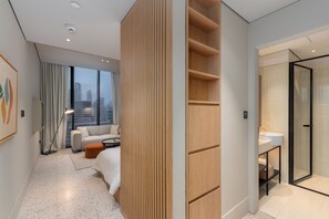 A modern hotel room with a city view, a sofa, a wooden panel divider, a glass shower, and a small table. - Burj View Studio Near Downtown and Canal (Dubai)