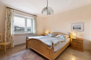 Un dormitorio con una cama de madera, una mesita de noche con una lámpara, una silla, una ventana con cortinas y una lámpara colgante.