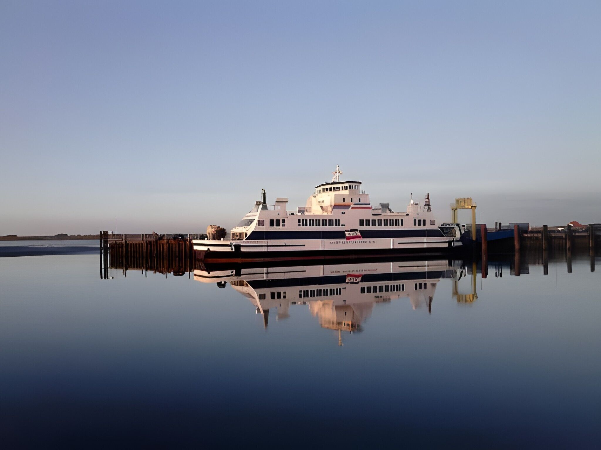 A large, stationary ferry docked at a pier with a clear reflection in the calm water.