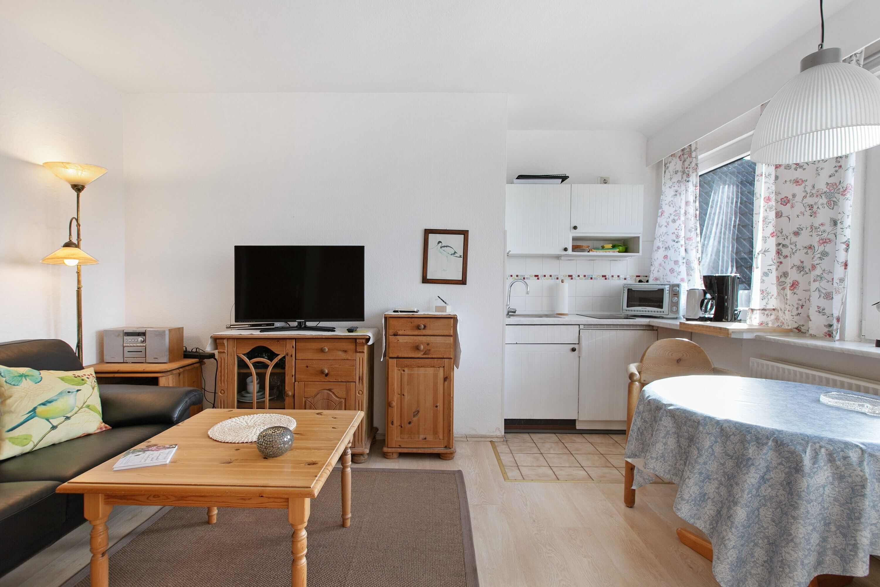 A compact living space with a sofa, a wooden table, a TV, a kitchen area with a microwave, and a dining table with a floral tablecloth.