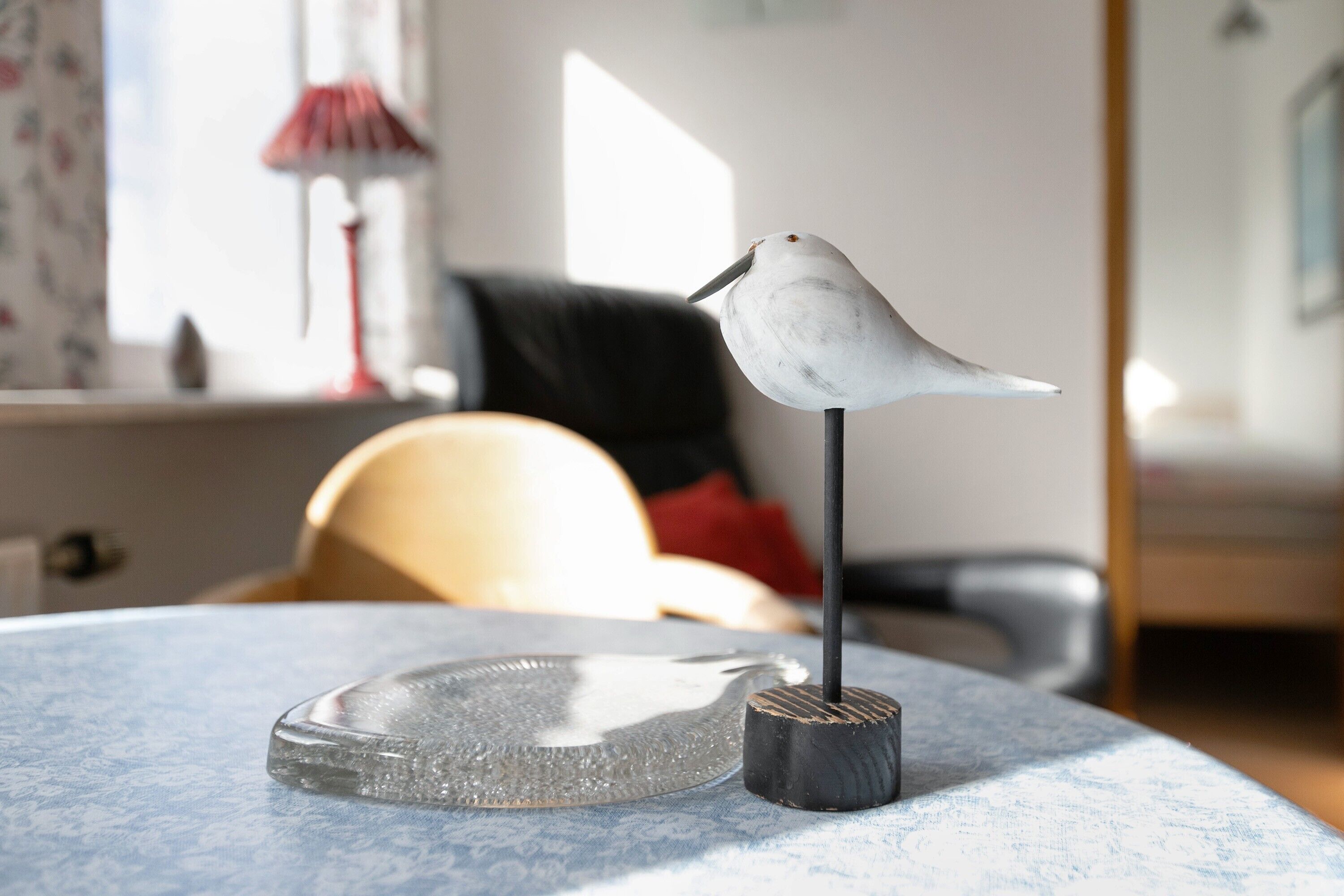 A bird figurine on a stand, a glass object, and a chair in a room with a lamp and a window.