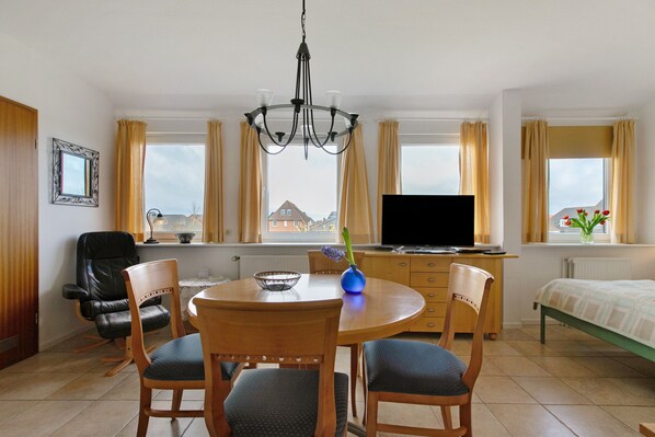 A room with a dining table, chairs, a TV, a bed, and windows with curtains.