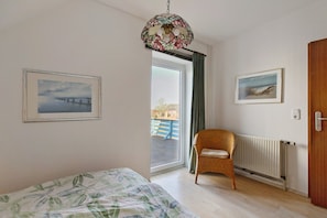 Une pièce comprenant un lit, une chaise en osier, un radiateur et deux tableaux encadrés accrochés au mur.