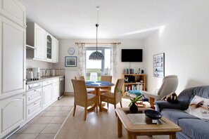 Une cuisine moderne avec des meubles blancs, un coin repas avec des chaises en osier, un téléviseur à écran plat et un canapé avec un coussin.