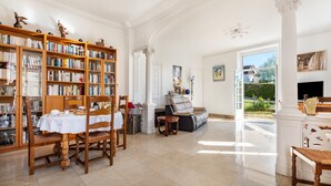 A spacious room with a large bookshelf, a dining area with a table and chairs, and a view through an open door to an outdoor garden. - Dentelles Guest Room (Neuvic)