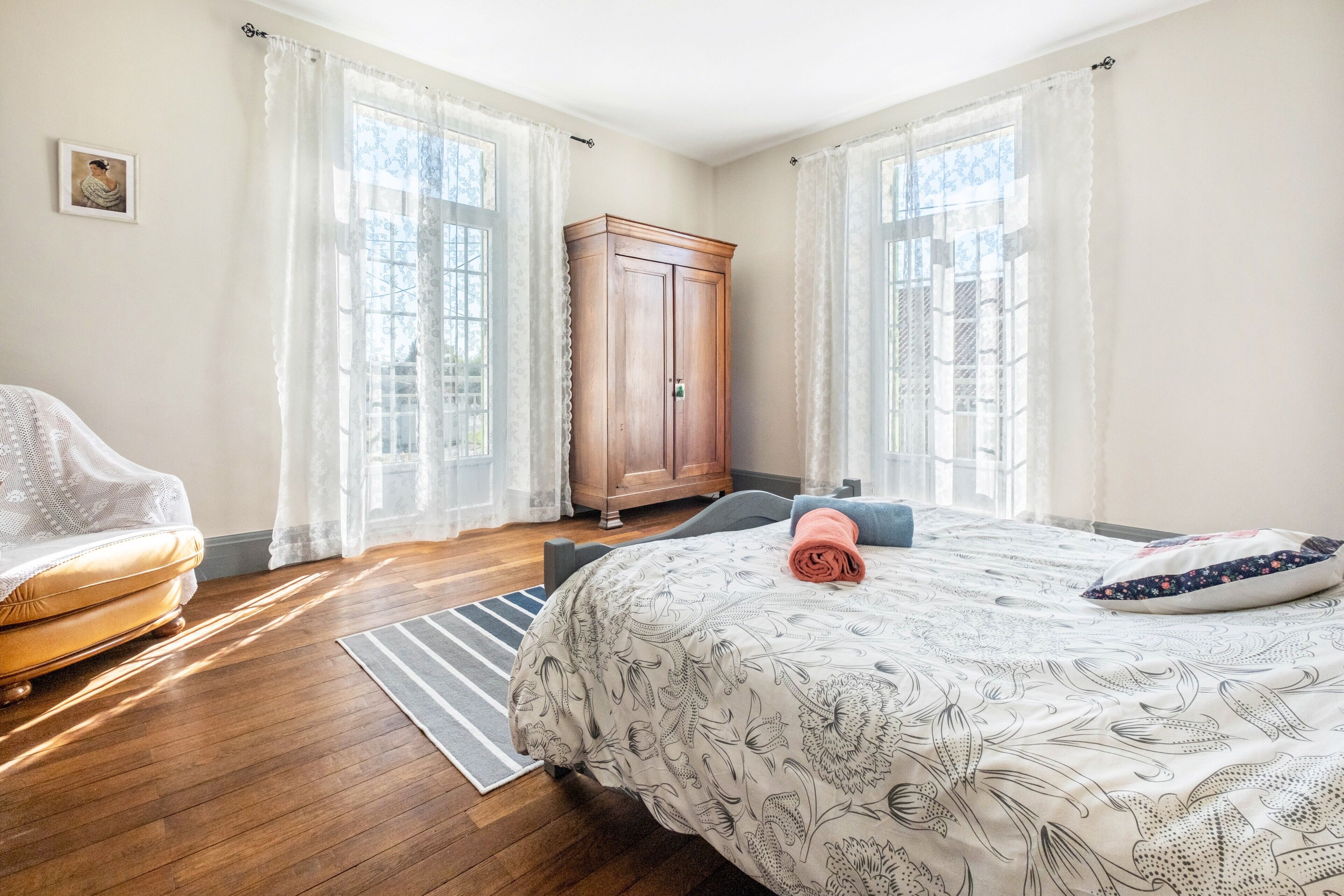 Une chambre à coucher avec un lit, une armoire en bois, une chaise et une fenêtre avec des rideaux.