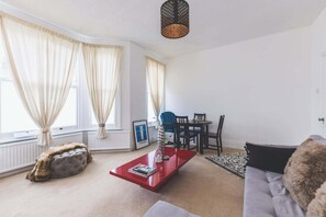 A living room with a red coffee table, a grey sofa, a blue chair, and a dining table with chairs. There are large windows with curtains, a radiator, and a hanging light fixture.