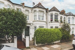 A row of white Victorian-style townhouses with bay windows, a hedge, and a car parked on the street.