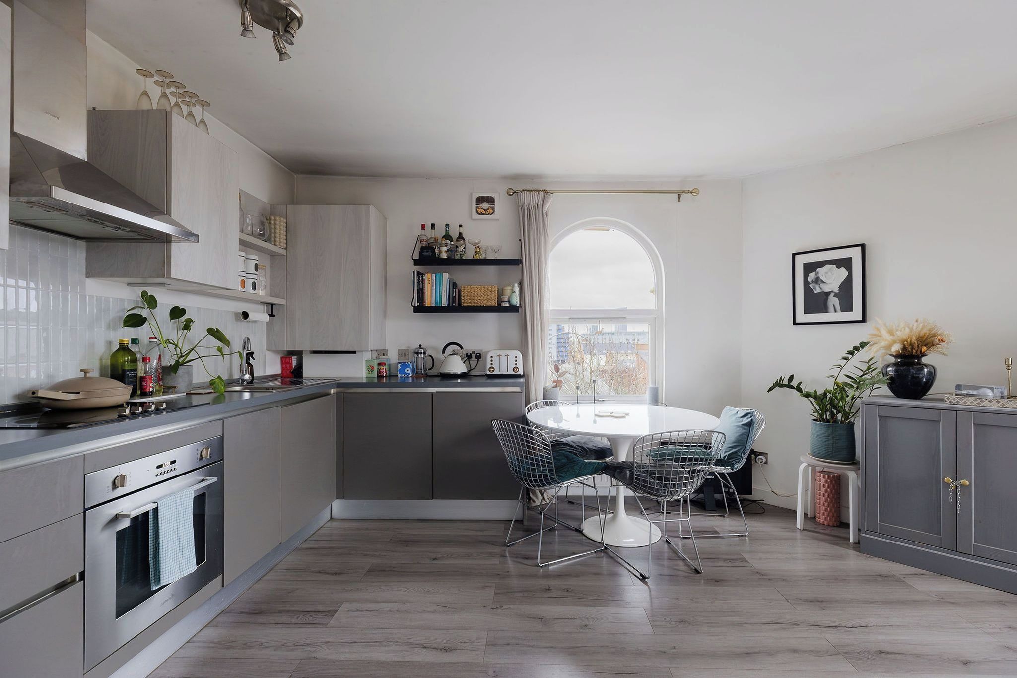 A modern kitchen with a dining area, featuring a built-in oven, a dining table with chairs, and a view through a window.
