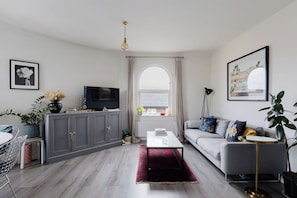 A modern living room with a grey sofa, a TV on a cabinet, a coffee table, and a large window with curtains. - Chic 1BD Flat in Restored English Pub, East London (Greater London)