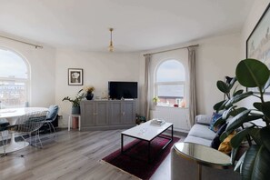 Een moderne woonkamer met een eetgedeelte, grijze kasten, een flatscreen televisie en grote ramen.