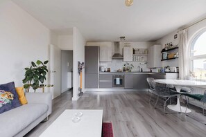 Une cuisine moderne avec des meubles gris, un plan de travail blanc et un coin repas comprenant une table ronde et des chaises en fil de fer.